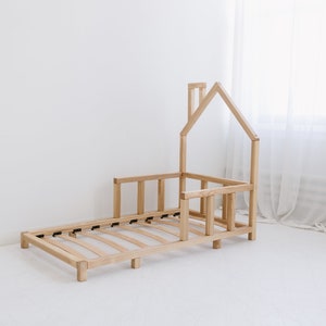 K&ouml;nnte beinhalten: Ein h&ouml;lzerner Kleinkindbettgestell in Form eines Hauses mit einem Lattenrost und einem Sicherheitsgel&auml;nder. Das Bettgestell ist aus Naturholz gefertigt und hat ein schlichtes, modernes Design.
