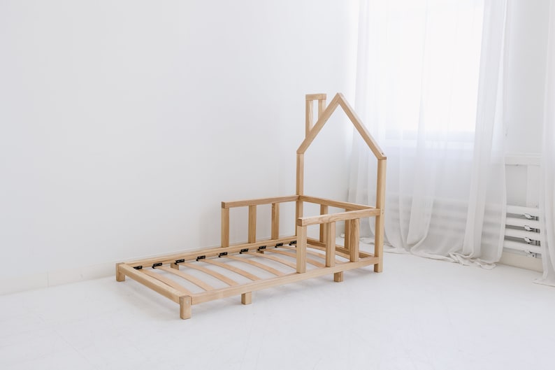 K&ouml;nnte beinhalten: Ein h&ouml;lzerner Kleinkindbettgestell in Form eines Hauses mit einem Lattenrost und einem Sicherheitsgel&auml;nder. Das Bettgestell ist aus Naturholz gefertigt und hat ein schlichtes, modernes Design.