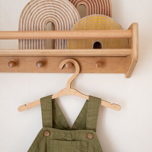 May include: A wooden wall-mounted shelf with three wooden rainbow decorations and a wooden hanger with a green overall hanging from it.