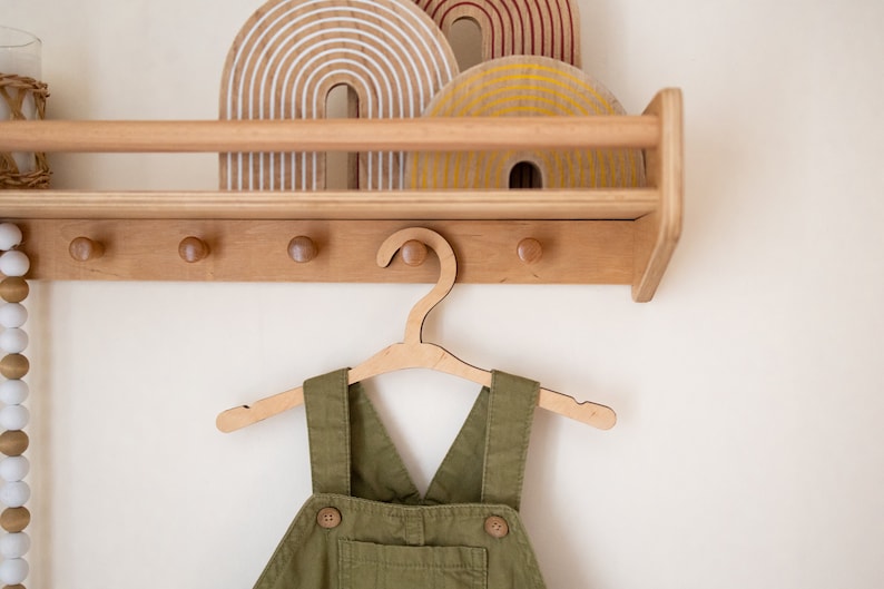 May include: A wooden wall-mounted shelf with three wooden rainbow decorations and a wooden hanger with a green overall hanging from it.