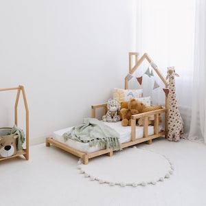 May include: A light wood-framed toddler bed with a house-shaped headboard is filled with stuffed animals and pillows. A green blanket is draped over the bed. A giraffe stuffed animal stands beside the bed. A round white rug with pom-pom trim is on the floor.