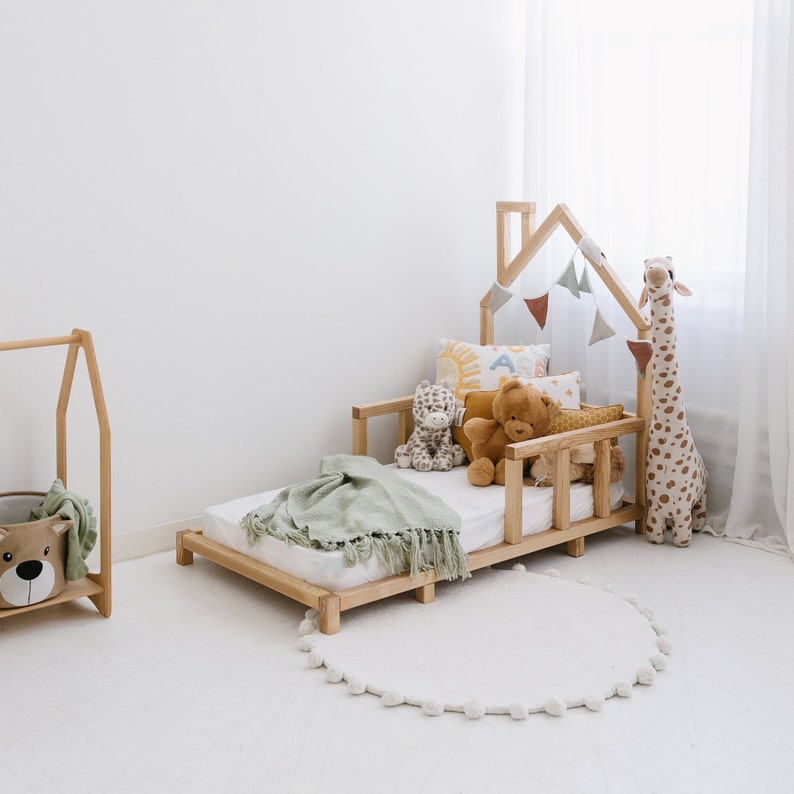 May include: A light wood-framed toddler bed with a house-shaped headboard is filled with stuffed animals and pillows. A green blanket is draped over the bed. A giraffe stuffed animal stands beside the bed. A round white rug with pom-pom trim is on the floor.