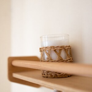 May include: A white candle in a glass jar with a woven rattan band sits on a wooden shelf with a beaded garland hanging below.