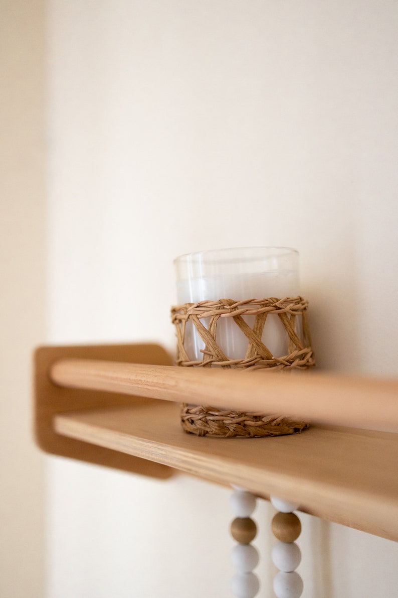 May include: A white candle in a glass jar with a woven rattan band sits on a wooden shelf with a beaded garland hanging below.