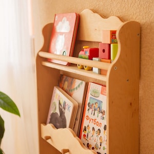 May include: A light-colored wooden bookshelf with a scalloped top, holding children's books and wooden toys. The books have colorful covers, and a small red toy train sits on a shelf. The bookshelf is against a light brown wall.