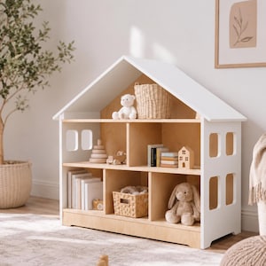 May include: A white and light wood dollhouse-shaped storage unit with a gabled roof and rectangular cutouts for windows. The interior features three levels of shelving divided into multiple compartments, containing books, woven baskets, and plush toys.