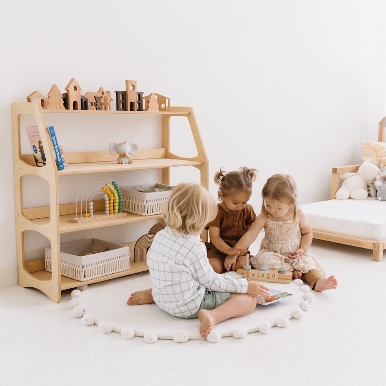 Op de afbeelding: Drie kinderen spelen met houten speelgoed op een wit tapijt voor een houten plank met speelgoed en boeken. De plank heeft drie planken en is gemaakt van licht hout. De kinderen zitten op een wit tapijt met een pompon rand. De kinderen spelen met een houten speelgoed dat cijfers erop heeft. De kinderen dragen casual kleding.