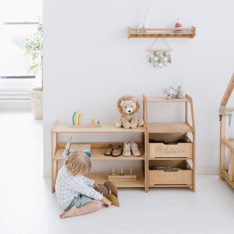 Pode incluir: Uma estante de madeira com duas prateleiras e uma gaveta. A gaveta &eacute; etiquetada com "Adam". A estante est&aacute; cheia de brinquedos, incluindo um le&atilde;o de pel&uacute;cia, um &aacute;baco arco-&iacute;ris e um conjunto de blocos de madeira. Uma crian&ccedil;a est&aacute; sentada no ch&atilde;o em frente &agrave; estante, brincando com um brinquedo.