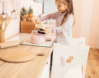 May include: A young girl in a striped shirt stands on a white learning tower and stirs a pink pot on a stovetop. The kitchen is decorated in pink and white.