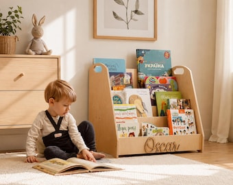 May include: A light-colored wooden bookshelf with three tiered shelves for displaying books. The front panel features the name Oscar in a cursive wooden cutout. The bookshelf has side handles and is filled with various children's books.