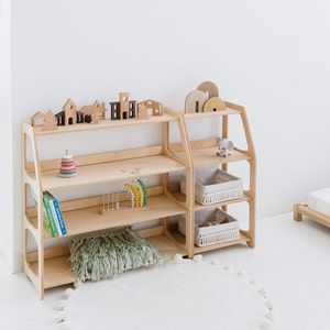 Op de afbeelding: Twee houten planken met elk drie planken. De bovenste plank heeft houten speelgoedhuisjes en een houten speelgoedboom. De tweede plank heeft een houten speelgoedabacus en een stapel houten ringen. De derde plank heeft een boek met de titel "Little Farm Friends". De onderste plank heeft een groen, pluche kussen. De planken zijn gemaakt van licht hout en hebben een natuurlijke afwerking.