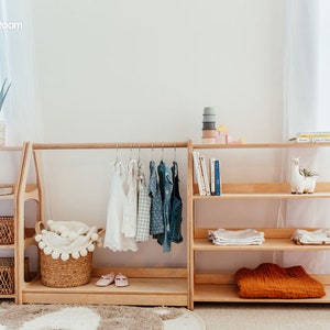 May include: A light-colored wooden modular storage unit featuring a central clothing rack with hangers, flanked by two shelving sections. The unit includes woven baskets, folded textiles, books, a small plant, and a llama-shaped decorative object.
