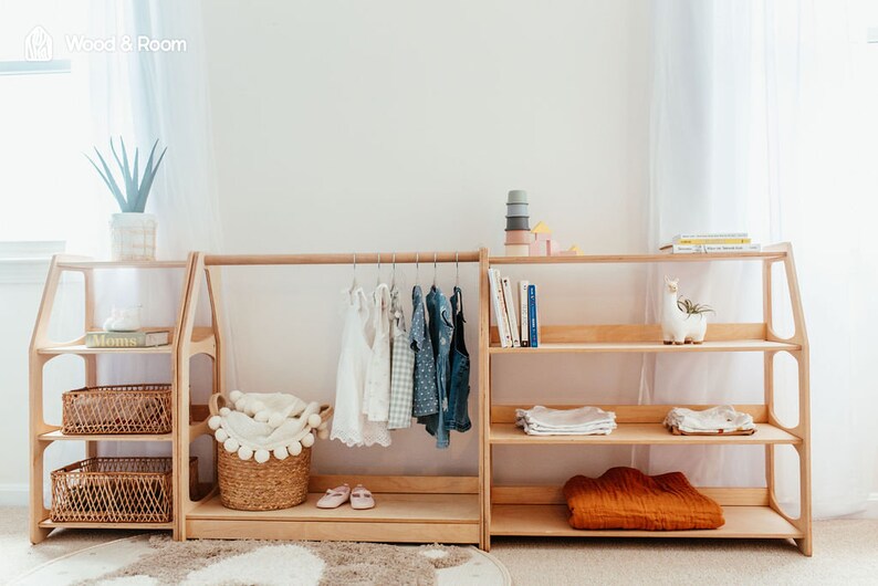 May include: A light-colored wooden modular storage unit featuring a central clothing rack with hangers, flanked by two shelving sections. The unit includes woven baskets, folded textiles, books, a small plant, and a llama-shaped decorative object.