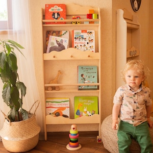 May include: A wooden bookshelf filled with colorful children's books, toys, and a wooden giraffe. The bookshelf is light-colored and has a cloud-shaped design. A young child is sitting nearby.