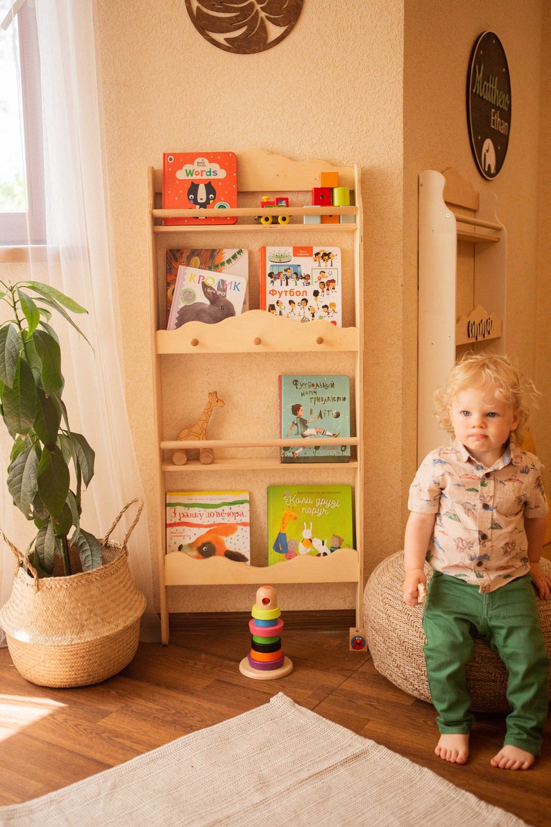 May include: A wooden bookshelf filled with colorful children's books, toys, and a wooden giraffe. The bookshelf is light-colored and has a cloud-shaped design. A young child is sitting nearby.