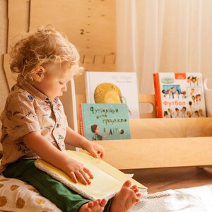 May include: A young child sits on a pillow and reads a book. The child is wearing a light brown shirt with a pattern of dinosaurs. The child is holding a yellow book with the pages open. Behind the child is a wooden bookshelf with several books on the shelves. The books have titles in Russian. The bookshelf is white and has a light wood finish.