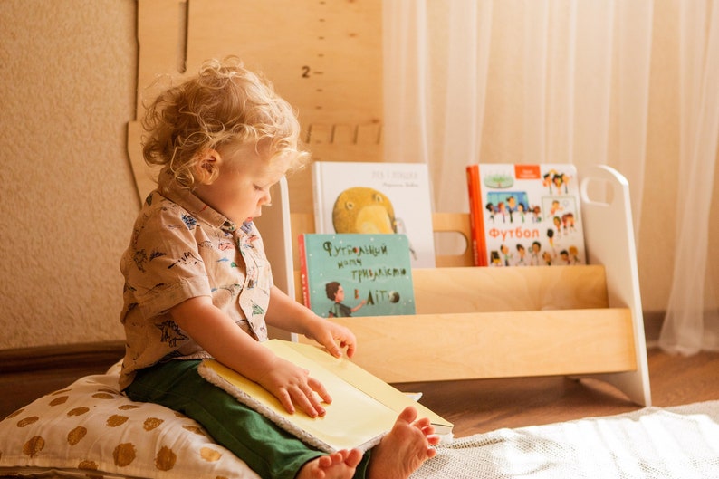 May include: A young child sits on a pillow and reads a book. The child is wearing a light brown shirt with a pattern of dinosaurs. The child is holding a yellow book with the pages open. Behind the child is a wooden bookshelf with several books on the shelves. The books have titles in Russian. The bookshelf is white and has a light wood finish.