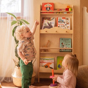 May include: Two children playing with a wooden toy stacking ring set. The girl on the left is reaching for a book on a wooden bookshelf with multiple shelves. The bookshelf is filled with colorful children's books. The books have titles like "Words", "Kromk", "Futbol", and "3 razy po 10 neubora".