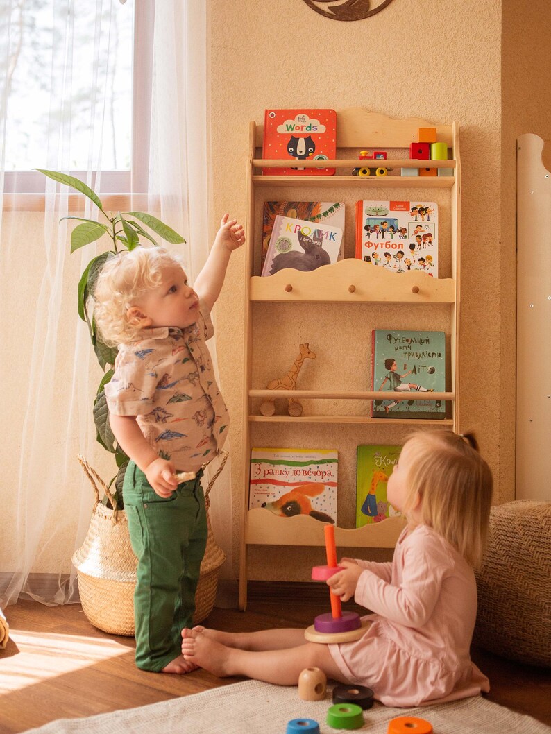 May include: Two children playing with a wooden toy stacking ring set. The girl on the left is reaching for a book on a wooden bookshelf with multiple shelves. The bookshelf is filled with colorful children's books. The books have titles like "Words", "Kromk", "Futbol", and "3 razy po 10 neubora".