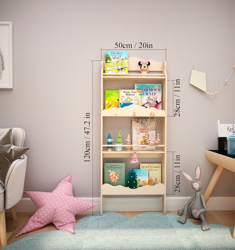 May include: A light-colored wooden bookshelf, 47.2 inches tall and 20 inches wide, with three shelves. The shelves hold children's books and decorative items. A pink star-shaped pillow and a gray stuffed rabbit are on the floor.