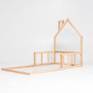 May include: A wooden bed frame shaped like a house with a chimney. The frame is made of unfinished wood and has a slatted headboard and footboard.