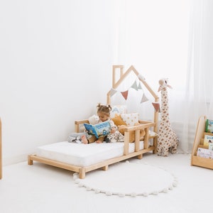 K&ouml;nnte beinhalten: Ein Kinderbett aus Holz mit einem hausf&ouml;rmigen Kopfteil und einer wei&szlig;en Matratze. Ein Kind sitzt auf dem Bett und liest ein Buch. Auf dem Bett liegen Stofftiere und Kissen. Im Hintergrund steht ein B&uuml;cherregal aus Holz mit B&uuml;chern.