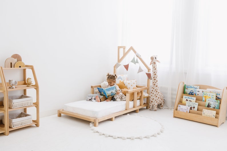 K&ouml;nnte beinhalten: Ein Kinderbett aus Holz mit einem hausf&ouml;rmigen Kopfteil und einer wei&szlig;en Matratze. Ein Kind sitzt auf dem Bett und liest ein Buch. Auf dem Bett liegen Stofftiere und Kissen. Im Hintergrund steht ein B&uuml;cherregal aus Holz mit B&uuml;chern.