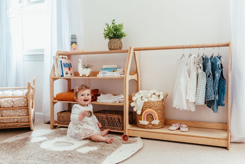 May include: A light wood children's room setup with a bookshelf, clothing rack, and a crib. The bookshelf holds books and a llama figurine. The clothing rack displays various children's clothes. A smiling baby sits on a rug.