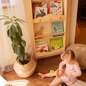 May include: A light wooden bookshelf filled with children's books, a toy train, and wooden blocks. A young child in a pink dress plays with a wooden stacking toy on a gray rug. A potted plant in a woven basket sits nearby.