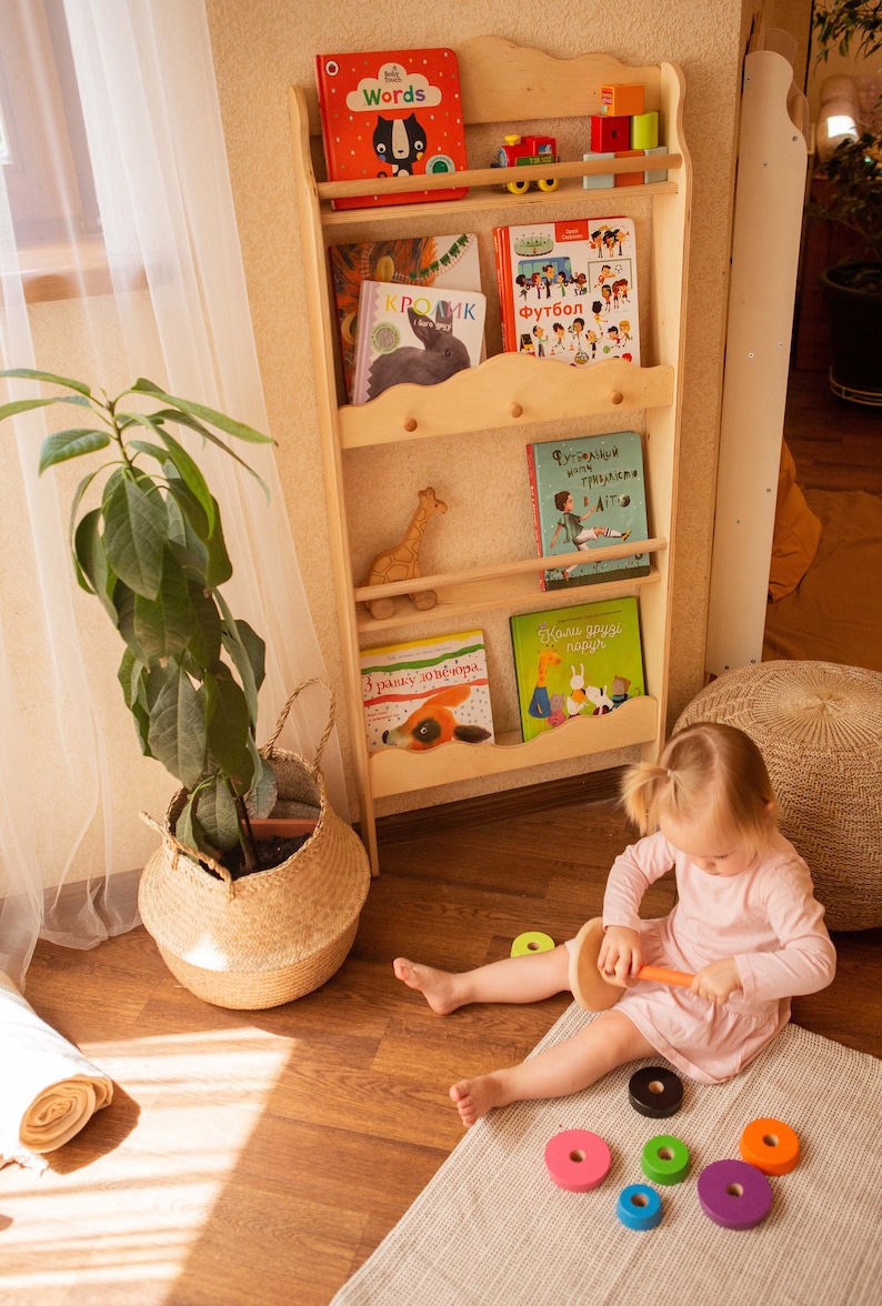 May include: A light wooden bookshelf filled with children's books, a toy train, and wooden blocks. A young child in a pink dress plays with a wooden stacking toy on a gray rug. A potted plant in a woven basket sits nearby.