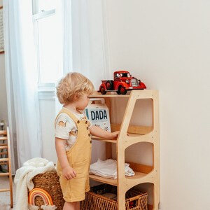 May include: A light wood, three-shelf storage unit with a red toy truck on top. A child in yellow overalls stands next to the unit. A woven basket with a rainbow decor and a rug with a paw print design are also visible.