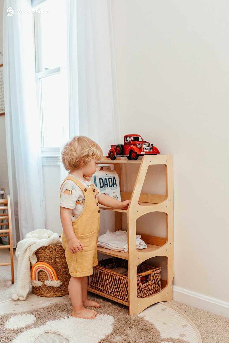 May include: A light wood, three-shelf storage unit with a red toy truck on top. A child in yellow overalls stands next to the unit. A woven basket with a rainbow decor and a rug with a paw print design are also visible.