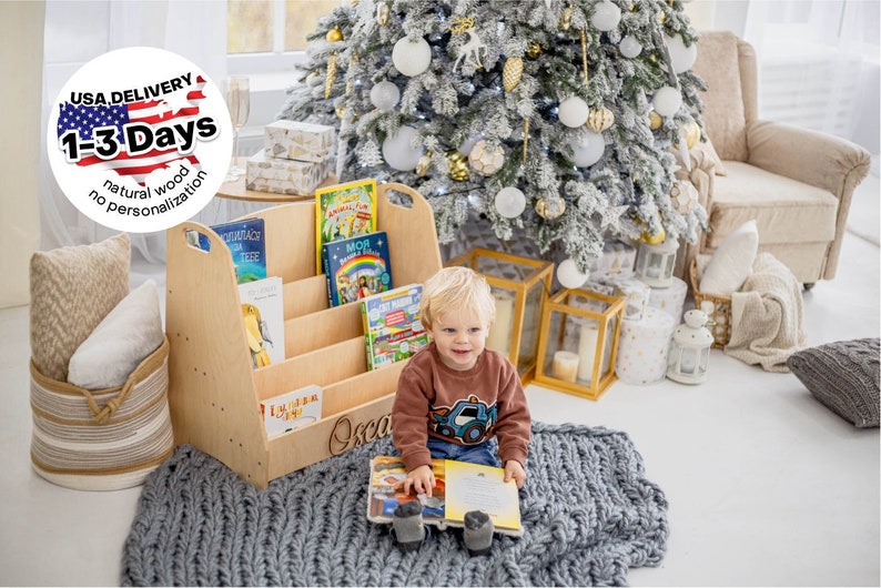 K&ouml;nnte beinhalten: Ein B&uuml;cherregal aus Holz mit vier Regalbrettern zum Aufbewahren von B&uuml;chern. Das B&uuml;cherregal ist mit dem Namen "Oscar" personalisiert. Ein kleines Kind sitzt auf einem grauen Strickteppich und liest ein Buch. Das B&uuml;cherregal steht vor einem Weihnachtsbaum, der mit wei&szlig;en und goldenen Kugeln geschm&uuml;ckt ist.