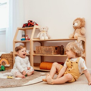 May include: A light-colored wooden toy storage shelf with multiple open levels, featuring a house-like frame design. The shelf holds a red toy truck, a white teddy bear, a lion plush, and a woven basket, with various toys scattered nearby.
