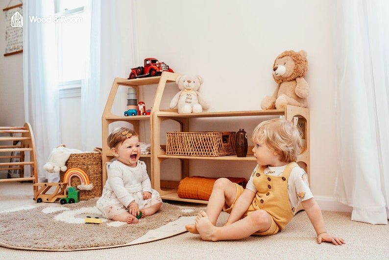 May include: A light-colored wooden toy storage shelf with multiple open levels, featuring a house-like frame design. The shelf holds a red toy truck, a white teddy bear, a lion plush, and a woven basket, with various toys scattered nearby.