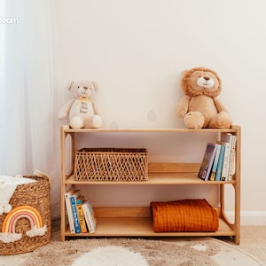 May include: A light-toned wooden bookshelf with two shelves, holding a woven basket, books, and a folded orange blanket. A beige teddy bear and a brown lion plush toy sit on top. A woven basket with a rainbow design sits to the left of the shelf.