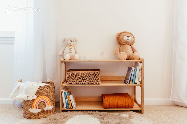 May include: A light-toned wooden bookshelf with two shelves, holding a woven basket, books, and a folded orange blanket. A beige teddy bear and a brown lion plush toy sit on top. A woven basket with a rainbow design sits to the left of the shelf.