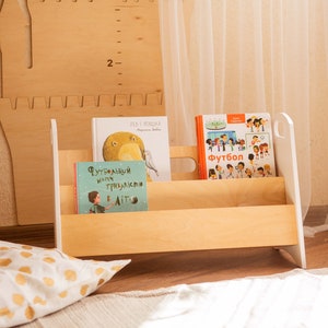 May include: A wooden bookshelf with three shelves, painted white, with a handle on the side. The bookshelf is holding three children's books, one with the title "Футбольный матч и триллизм" and another with the title "Футбол".