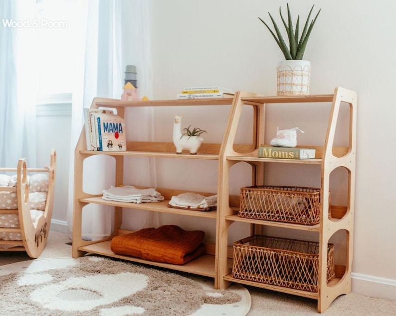 K&ouml;nnte beinhalten: Ein helles Holzregal mit mehreren offenen F&auml;chern, bestehend aus einem hohen und einem niedrigeren Seitenteil. Die Regale enthalten B&uuml;cher mit den Titeln MAMA und Moms, eine wei&szlig;e Lama-Figur, zwei geflochtene K&ouml;rbe und eine gr&uuml;ne Topfpflanze.