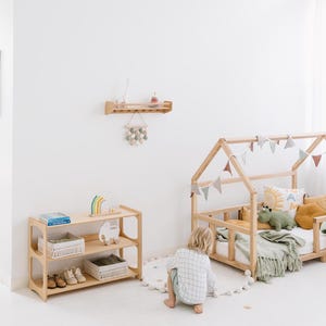 Pode incluir: Uma estrutura de cama de madeira em forma de casa com um cobertor verde e branco e um animal de pel&uacute;cia. Uma crian&ccedil;a est&aacute; sentada no ch&atilde;o em frente &agrave; cama. H&aacute; uma prateleira de madeira na parede com uma figura de girafa de madeira e um caminh&atilde;o de brinquedo de madeira em uma prateleira abaixo.
