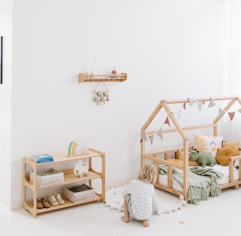 Pode incluir: Uma estrutura de cama de madeira em forma de casa com um cobertor verde e branco e um animal de pel&uacute;cia. Uma crian&ccedil;a est&aacute; sentada no ch&atilde;o em frente &agrave; cama. H&aacute; uma prateleira de madeira na parede com uma figura de girafa de madeira e um caminh&atilde;o de brinquedo de madeira em uma prateleira abaixo.