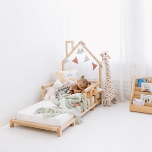May include: A light wood-framed toddler bed with a house-shaped frame and a small child lying on it. The bed is decorated with a green blanket, stuffed animals, and a banner of pastel-colored flags. A tall giraffe plush toy stands nearby.