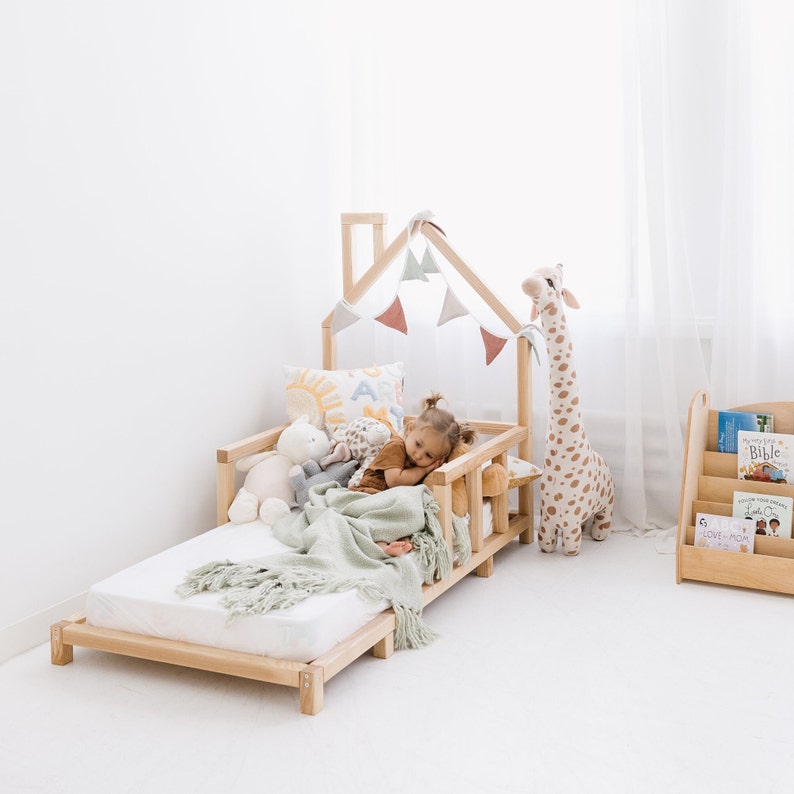 May include: A light wood-framed toddler bed with a house-shaped frame and a small child lying on it. The bed is decorated with a green blanket, stuffed animals, and a banner of pastel-colored flags. A tall giraffe plush toy stands nearby.