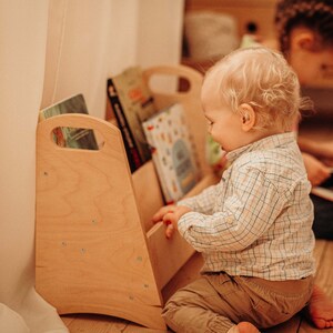 Puede incluir: Una estanter&iacute;a de libros para ni&ntilde;os de madera clara con bordes redondeados y un asa a cada lado. Dos ni&ntilde;os est&aacute;n sentados en el suelo junto a la estanter&iacute;a, mirando libros.