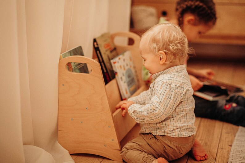 Puede incluir: Una estanter&iacute;a de libros para ni&ntilde;os de madera clara con bordes redondeados y un asa a cada lado. Dos ni&ntilde;os est&aacute;n sentados en el suelo junto a la estanter&iacute;a, mirando libros.