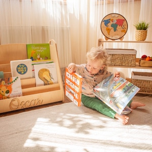 May include: A wooden bookshelf with the name "Oscar" engraved on the front. The bookshelf is filled with children's books and is sitting on a white rug. A young child is sitting on the rug and reading a book.