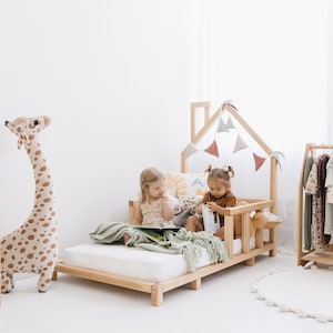 May include: A light wood-framed children's bed with a house-shaped frame and decorative bunting. Two young children are sitting on the bed, reading. A large stuffed giraffe and a clothing rack are also in the room.