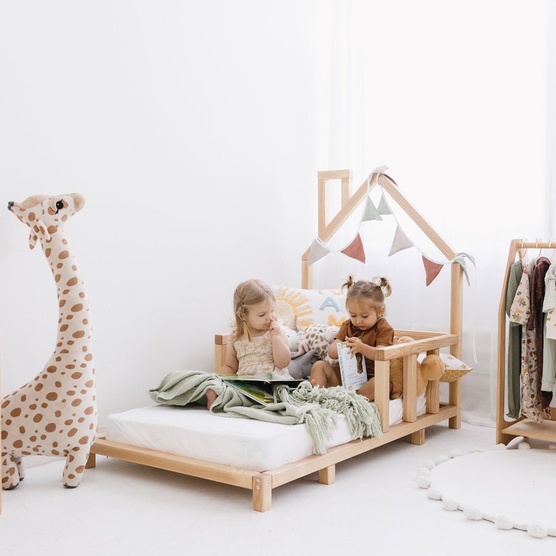 May include: A light wood-framed children's bed with a house-shaped frame and decorative bunting. Two young children are sitting on the bed, reading. A large stuffed giraffe and a clothing rack are also in the room.