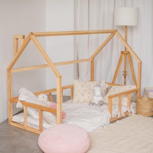 May include: A wooden house-shaped bed frame with a white mattress and pillows. The bed is decorated with stuffed animals and a pink throw pillow. The bed is in a room with a white rug and a wooden floor.