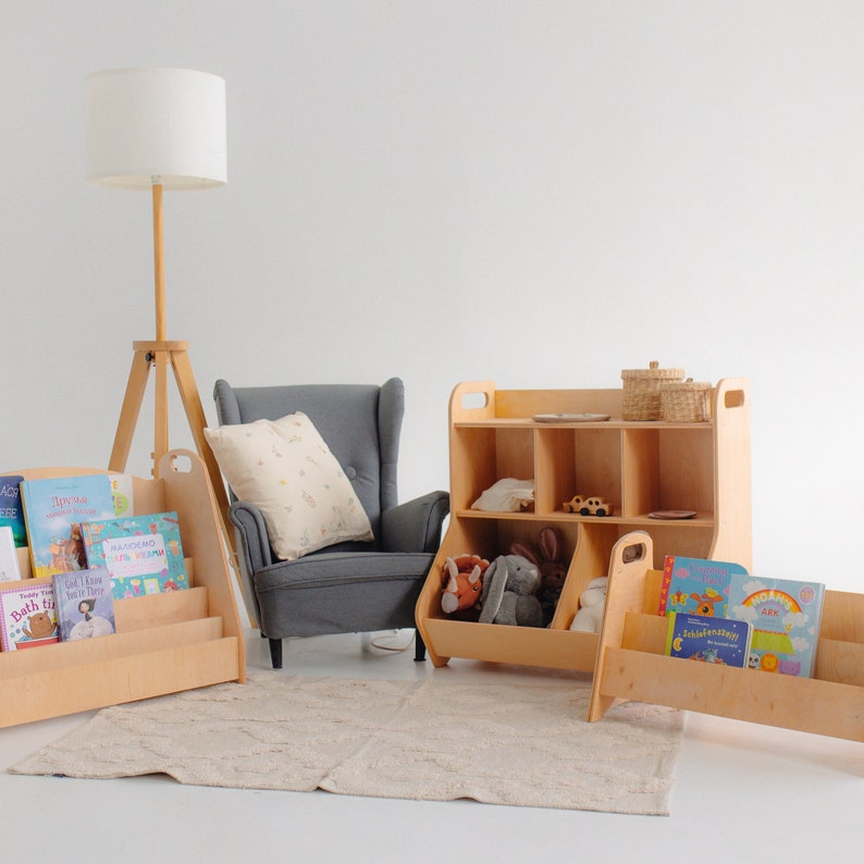 K&ouml;nnte beinhalten: Ein Satz Holzregale mit einem grauen Sessel und einer Stehlampe. Die Regale haben offene F&auml;cher und einen Spielzeugbeh&auml;lter. Die Regale sind mit B&uuml;chern und Spielzeug gef&uuml;llt.